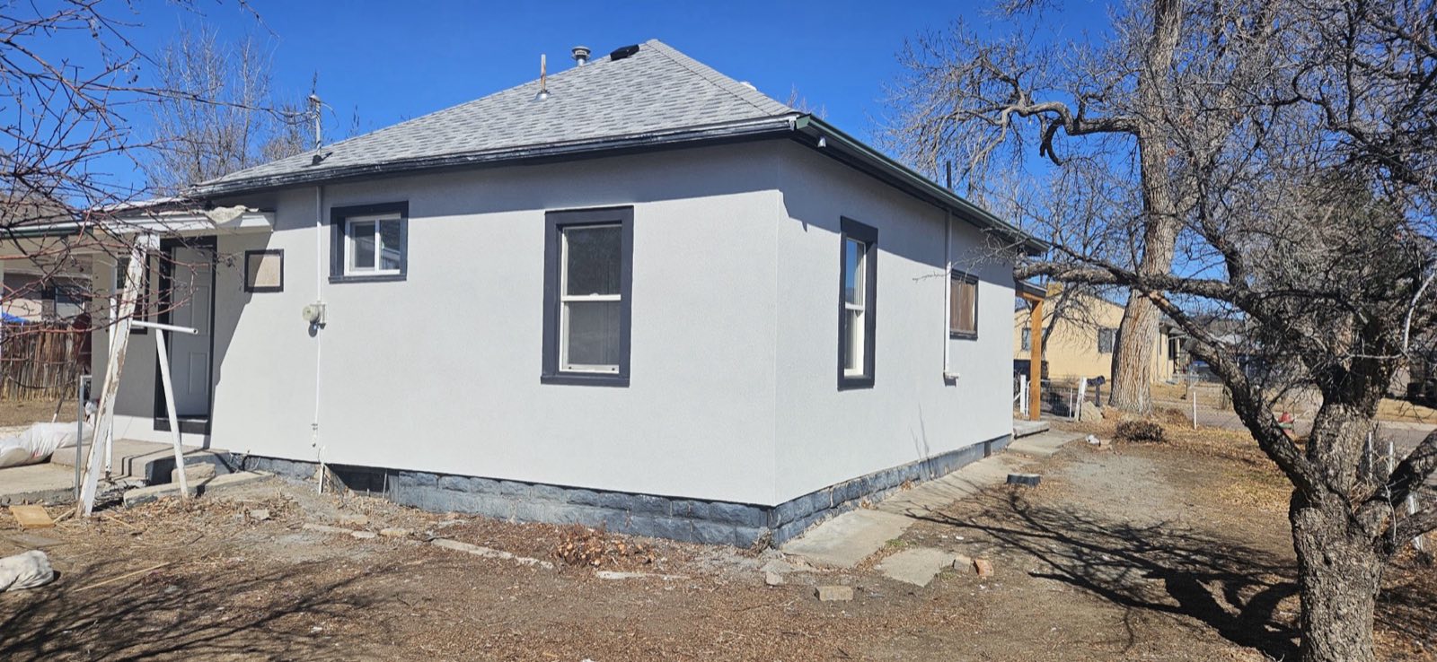 A finished house in pale gray stucco, clean to the foundation line, bare trees behind it.