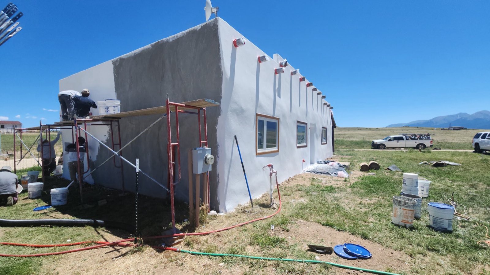 A long single-story building part-way through a new stucco coat, one of the crew working from a plank scaffold, buckets and hoses on the grass, the mountains behind.